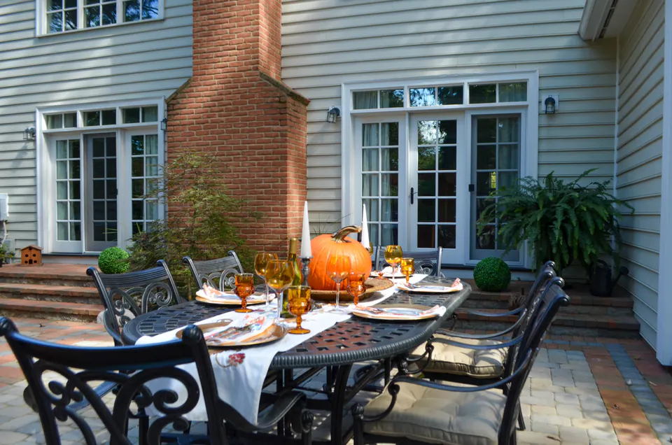 Outdoor dining table beside house doors with brick chimney, paver patio, pumpkins, and fall table setting.